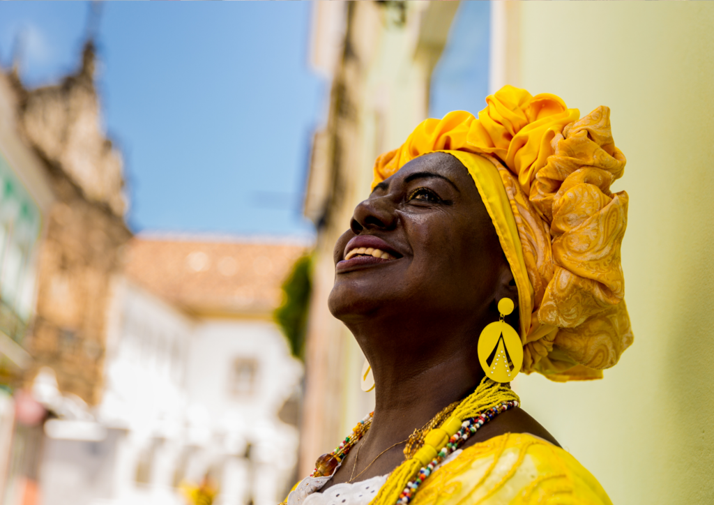 Een portretfoto van een Afro-Braziliaanse vrouw in traditionele felgele kleding, die omhoog kijkt en glimlacht, met een onscherpe historische achtergrond.