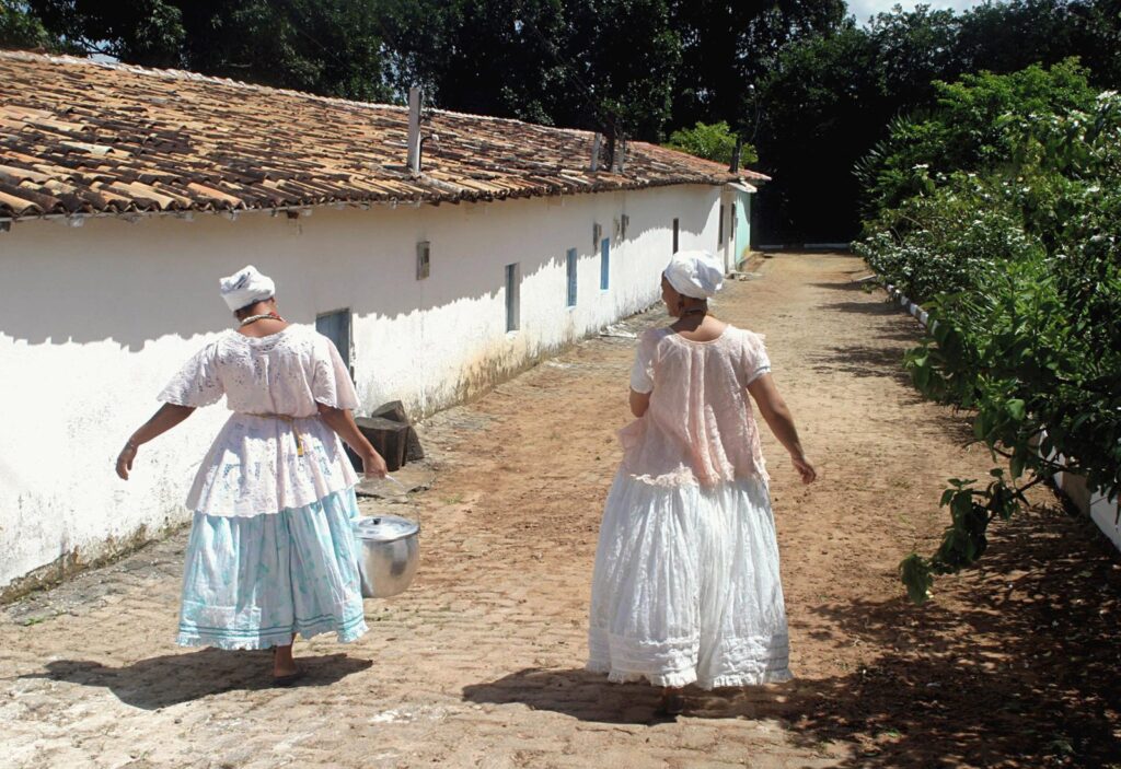 Twee vrouwen (Baianas) in traditionele Candomblé-kleding lopen over het terrein van een terreiro in Brazilië. Ze dragen witte hoofddoeken en wijde, ceremoniële rokken, terwijl een van hen een metalen kookpan draagt, wat wijst op de voorbereiding van een ritueel of gemeenschappelijke maaltijd.