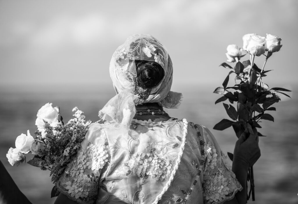 Een vrouw van achteren gefotografeerd in traditionele Afro-Braziliaanse kleding, met een kanten hoofddoek en boeketten witte rozen, uitkijkend over de zee tijdens een religieus eerbetoon (voor de Orixá Yemanjá).