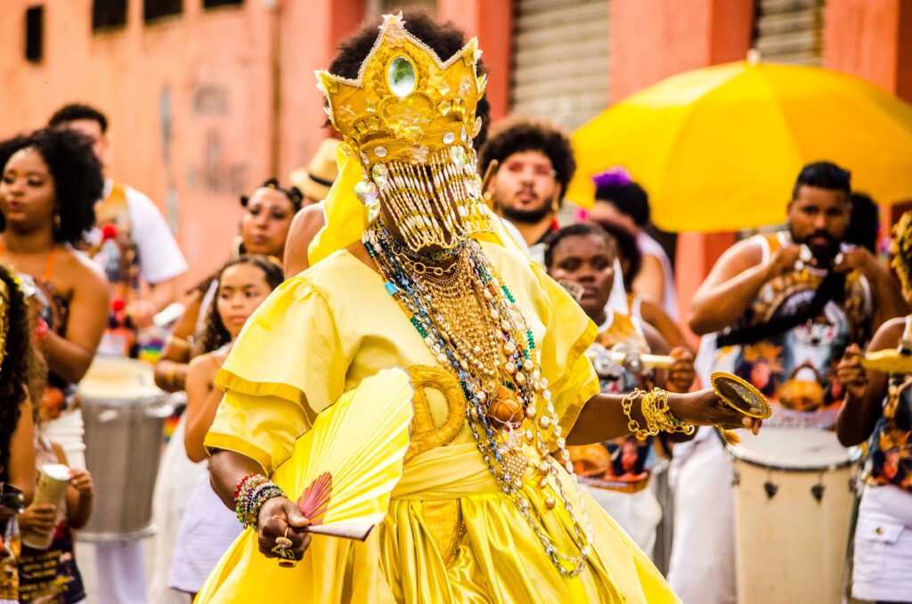 Een persoon in een weelderige, goudgele jurk en een gouden kroon met een kralensluier, die de Afro-Braziliaanse godheid Oxum uitbeeldt tijdens een straatfeest.