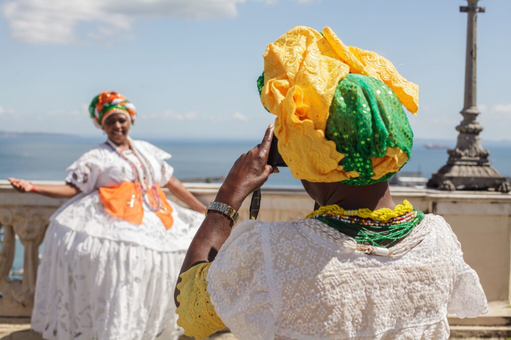 Twee traditionele Baianas in Salvador, Bahia, waarvan de een een foto maakt van de ander die poseert bij een uitkijkpunt. Beiden dragen de karakteristieke wijde kanten jurken, kleurrijke kralenkettingen en kunstig geknoopte hoofddoeken (torços) in felle kleuren zoals geel, groen en oranje, tegen de achtergrond van de oceaan.