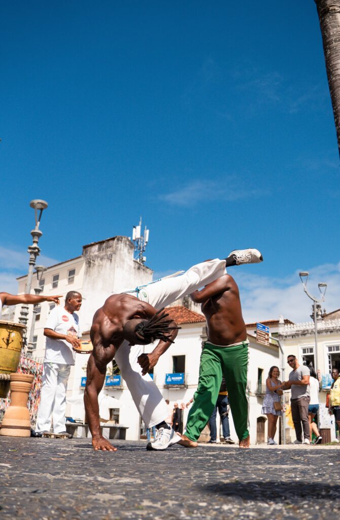 Mannen die capoeira beoefenen op een historisch, met kasseien bestraat plein in Brazilië onder een strakblauwe lucht.